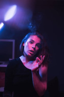 Parlak makyajlı ve yüzünde parıltı olan güzel bir kadın neon renkli bir ışıkla bir odada dikiliyor ve kameraya bakıyor. Dikey.