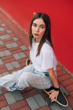 Beton kaldırımda kaykayıyla oturan çekici bir kaykaycının dikey portresi. Arka planda kırmızı duvar.