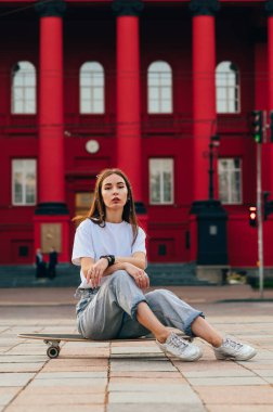 Kaykayında oturan, fotoğraf için poz veren ve kırmızı bir binanın önündeki kameraya bakan rahat ve genç bir bayan patencinin dikey çekimi..