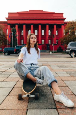 Vertical photo of a stylish skater woman in baggy jeans and white T-shirt sitting on a skateboard, looking away. City street and red building on a background. clipart