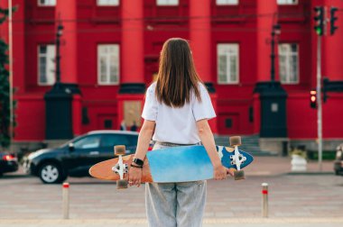 Kırmızı binanın önündeki bir caddede elinde renkli kaykayıyla günlük giysiler içinde duran bir kadın kaykaycının arka görüntüsü..