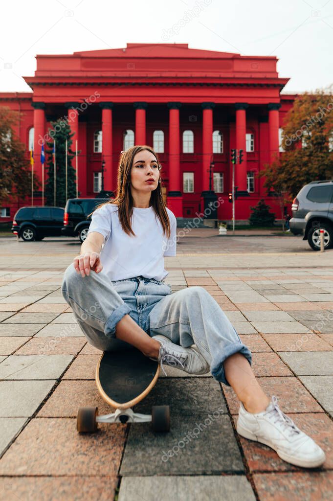 Vertical photo of a stylish skater woman in baggy jeans and white T-shirt sitting on a skateboard, looking away. City street and red building on a background.