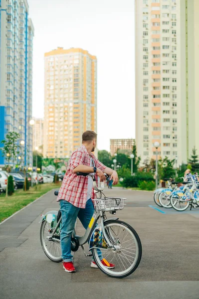 Man with a rented bicycle stands in the background of an area with high-rise buildings and looks back. Walking around the yard on a sharing bike