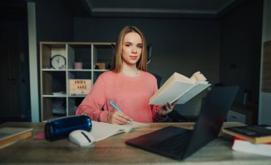 Evde dizüstü bilgisayarın yanında oturan sevimli bir öğrenci elinde bir kitapla kameraya bakıyor yüzünde bir gülümsemeyle, defterine kalemle yazıyor. Çevrimiçi öğrenme