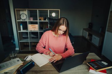 Genç bayan evde uzaktan ders çalışıyor, dizüstü bilgisayarla masaya oturuyor ve kalemle deftere yazıyor. Ev ödevi yapan kız öğrenci.