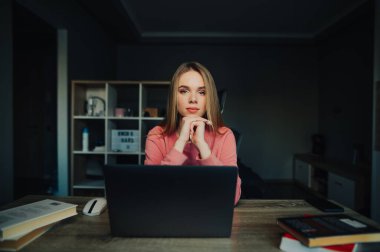 Çekici bir kız öğrenci evinde bir dizüstü bilgisayar, kitaplar ve bir defterle masada oturuyor ve kameraya poz veriyor. Evde ders çalışmak..