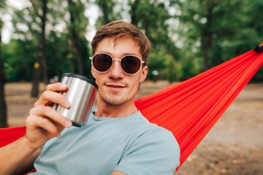 Elinde bir bardak içkiyle ormanda dinlenen ve kameraya bakıp gülümseyen genç bir adamın portresi. Resmi kapat.