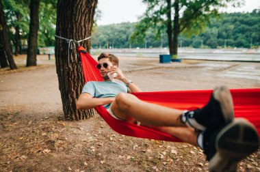 Konsantre genç adam parkta dinleniyor hamakta yatıyor, başka tarafa bakıyor ve telefonda konuşuyor. Adam parkta bir hamakta uzanıyor ve telefonla arıyor..