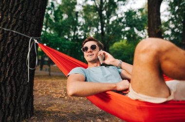 Güneş gözlüklü neşeli bir genç adam ağaçtaki bir hamakta dinleniyor ve telefonda gülümsüyor..