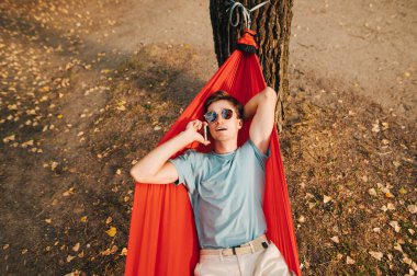 Hafif gündelik giysiler içindeki yakışıklı genç adam ormanda bir hamakta uzanıyor, dinleniyor ve telefonda konuşuyor. Turist, ormanda hamakta duran bir cep telefonu kullanıyor..