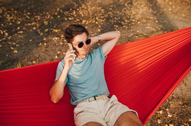 Konsantre genç adam parkta dinleniyor hamakta yatıyor, başka tarafa bakıyor ve telefonda konuşuyor. Adam parkta bir hamakta uzanıyor ve telefonla arıyor..