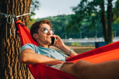Güneş gözlüklü genç adam gün batımında parkta bir hamakta uzanıyor, gülümsüyor ve telefonda konuşuyor..