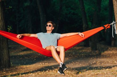 Güneş gözlüklü yakışıklı genç adam akşamları kırmızı bir hamakta oturur ve ciddi bir yüz ifadesiyle dinlenir..