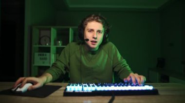 Kulaklık takmış, yüzü gülücükle video oyunları oynayan mutlu genç bir adam yeşil bir iç mekanı olan bir odada kameraya bakıyor ve yüzünde bir gülümsemeyle zaferle seviniyor.