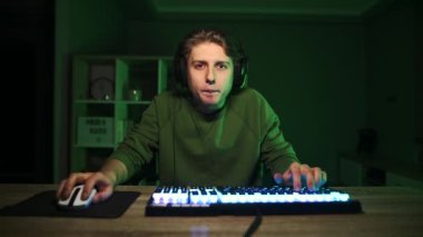 Yeşil ışıkları olan bir masada oturan ve ekranda gülümseyen bir gülümsemeyle bilgisayar oyunu oynayan sıradan giyinmiş ve kulaklık takan yakışıklı bir genç..