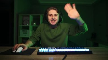 Neşeli oyuncu evde bilgisayar başında oturur ve çevrimiçi oyunlar izler, kameraya bakar ve avucuyla selam verir ve gülümser ve başparmağını kaldırır. Oyun.