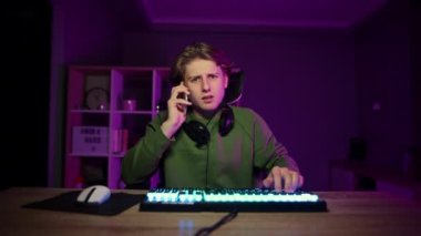 Boynuna kulaklık takmış yakışıklı bir adam mor ışığı olan bir odada bilgisayarın başında oturuyor ve telefonla konuşuyor.