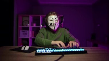 Yüzünde isimsiz bir maske ve kafasında kulaklık olan bir adam bilgisayar kullanıyor ve kameraya bir heavy metal hareketi gösteriyor..