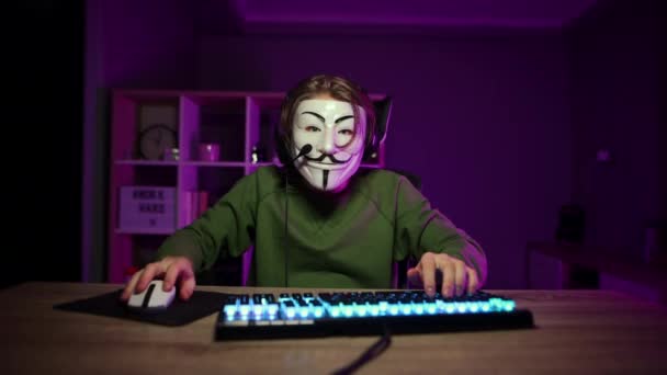 彼の顔とヘッドセット上の匿名のマスクで感情的なハッカーは自宅で夜コンピュータを使用し 彼の指を上げるアイデアのジェスチャーを示しています — ストック動画