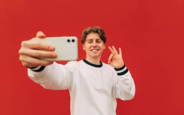 Beyaz tişörtlü yakışıklı adam kırmızı duvar arka planında selfie çekiyor ve tamam jest, şaşı ve akıllı telefon kamerasına bakıyor..