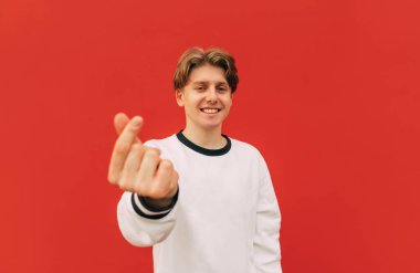 Kırmızı arka planda beyaz giysili neşeli yakışıklı genç adam başparmaklarını kaldırıyor kalp hareketi yapıyor ve kameraya bakıp gülümsüyor..