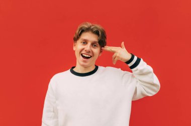 Çılgın bir gülüşü olan komik bir adam kırmızı arka planda duruyor ve parmağını şakağına dayayıp ateş ediyor, kameraya bakıyor ve gülüyor..