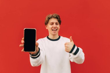 Neşeli genç adam baş parmağını gösteriyor ve kırmızı duvarda kamera var, gülümsüyor ve kameraya bakıyor..