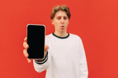 Beyaz kazaklı genç adam kırmızı bir duvarın arka planında duruyor ve kamerasında siyah bir erkan olan akıllı telefonu, yakın bir fotoğrafı gösteriyor. Boşluk ekranını kopyala.