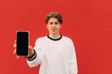 Ciddi yüzlü komik bir adam kırmızı arka planda duruyor ve kameraya siyah ekranlı bir akıllı telefon gösteriyor..
