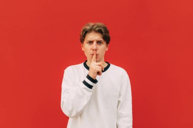 Beyaz kazaklı, ciddi yakışıklı bir adam kırmızı bir duvarın arkasında duruyor, kameraya bakıyor ve bir sessizlik hareketi gösteriyor..