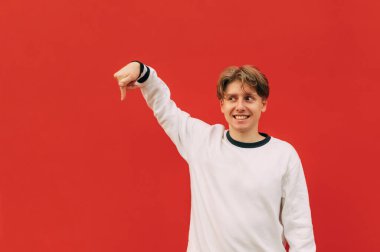 Yüzünde bir gülümseme olan komik adam parmağını aşağı ve arka planda beyaz bir kazak giyen kırmızı bir duvarı işaret ediyor..