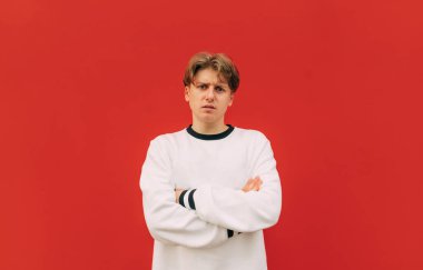 Hoşnutsuz bir gencin portresi kırmızı bir duvarın arkasında duruyor ve beyaz bir kazak giyen ciddi bir yüzle kameraya bakıyor..