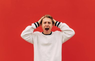 Beyaz kazaklı, şok olmuş bir adam kırmızı arka planda kameraya bakarak başını tutup bağırıyor. İzole edilmiş.