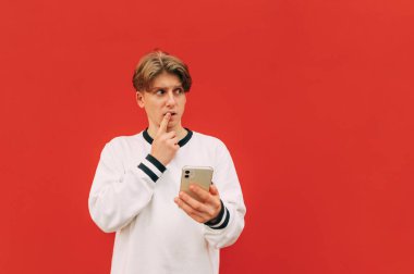 Beyaz kazaklı, düşünceli genç bir adamın portresi elinde bir akıllı telefonla kırmızı bir duvarın arka planında duruyor, fotokopi alanına bakıyor ve elini dudaklarına değdirdiğini düşünüyor.