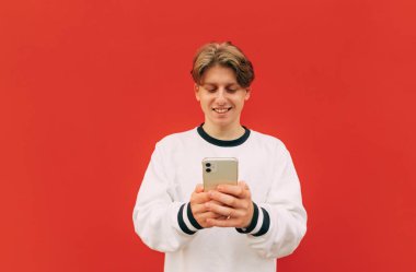 Beyaz tişörtlü pozitif adam kırmızı arka planda akıllı telefon kullanıyor ve gülümsüyor..