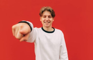 Mutlu genç adam, gündelik giysiler içinde parmağını kameraya gösteriyor. Yüzünde kırmızı bir tebessüm var..