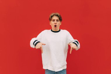 Şık giyinmiş, şık bir genç adam kırmızı duvarda şok olmuş bir yüzle kameraya bakar..