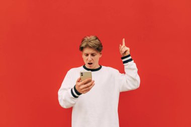 Şaşırmış bir adam kırmızı bir duvarın arka planında akıllı telefon kullanıyor ve reklam için parmağını yukarı kaldırıyor..