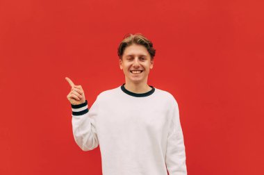 Kırmızı duvarın arkasında sıradan giyinmiş mutlu genç adam boş bir koltukta parmağını gösteriyor ve kameraya bakarken gülümsüyor..