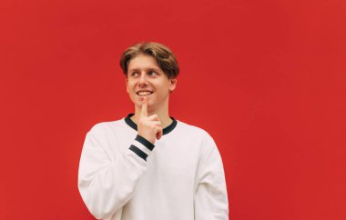 Yüzünde bir gülümseme olan düşünceli genç bir adam kırmızı bir arka planda duruyor ve mutlu bir yüzle başka yere bakıyor..