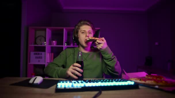 ヘッドフォンの若者は コンピュータの自宅で夜に座って 画面を見ながらピザやドリンクビールを食べています ゲーマーはジャンクフードで食事 — ストック動画