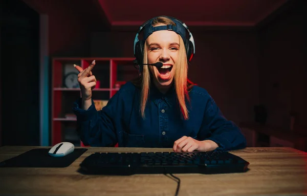 Happy female streamer in headset playing video games at home on computer looks at camera with happy face and shows gesture \