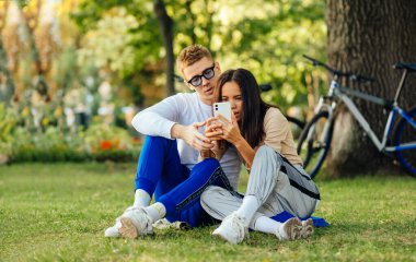 Genç modern çift parkta otururken mobil teknoloji kullanıyor. Bir kadın akıllı telefonuyla fotoğraf çekiyor, bir adam telefona bakıyor ve bir kadınla konuşuyor. Arka planda motorlar.