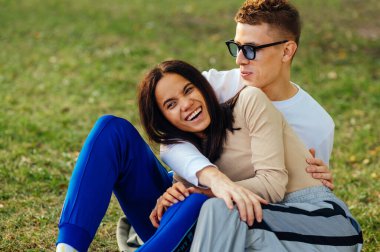 Genç ve güzel İspanyol kadın parkta erkek arkadaşıyla gülüyor ve sarılıyor yeşil çimlerin üzerinde oturuyor. Çift olumlu ve mutlu hissediyor..