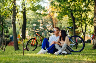 Konsantre olmuş birkaç bisikletçi şehir parkında dinleniyor, bir çimenlikte oturuyor ve ileriye bakıyor. El ele tutuşup sarılıyorlar..