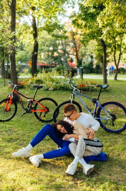 Şehir parkında yeşil bir çimenlikte otururken gülen ve sarılan neşeli bir çiftin dikey çekimi. Arka planda bisikletleri var. İlişki kavramı.