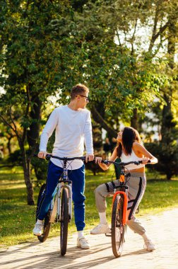 Gülen ve neşeli bir çiftin dikey çekimi renkli bisikletlerle şehir parkında bisiklet sürerken eğlenirken..
