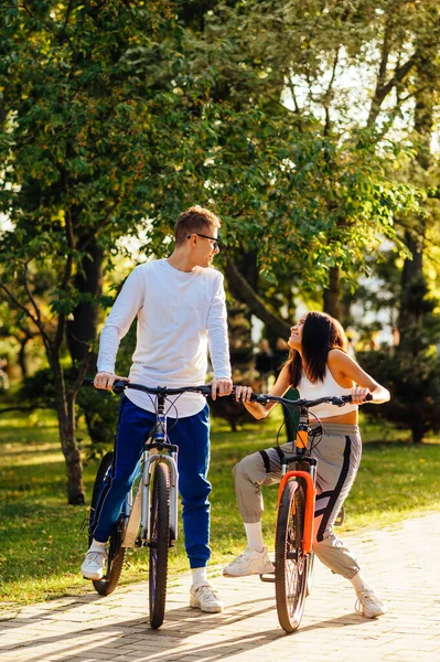 Gülen ve neşeli bir çiftin dikey çekimi renkli bisikletlerle şehir parkında bisiklet sürerken eğlenirken..