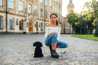 Yazlık kıyafetleriyle sokakta oturan, elinde sevimli siyah bir köpekle kameraya bakan bir kadın. Parkta evcil bir hayvanla vakit geçirmek..