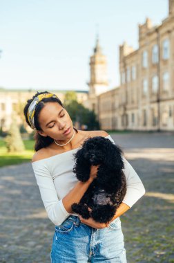 Güzel bir kadın portresi, gündelik kıyafetler içinde elinde oyuncak bir köpekle sokakta duran güzel bir manzaranın arka planında duruyor..